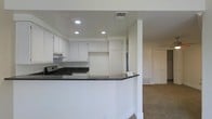 This single-story space showcases an open-concept layout that seamlessly connects the kitchen to the adjacent living area. The kitchen is outfitted with crisp white cabinetry that extends to the upper walls, offering generous storage. Dark granite countertops provide a striking contrast against the white surfaces, running along the perimeter and wrapping around a peninsula that serves as a natural divider between the kitchen and living space. A gas range and under-cabinet range hood are integrated into the design, and recessed lighting in the tray ceiling illuminates the kitchen efficiently. The peninsula's clean, curved edge adds a subtle architectural detail to the transition between spaces. The adjoining living area features neutral carpet flooring, providing a soft contrast to the hard surfaces in the kitchen. White walls throughout maintain a bright, cohesive aesthetic, while multiple ceiling vents suggest a well-distributed HVAC system. A door visible in the background hints at additional rooms or storage beyond the main living space. Overall, the layout is practical and functional, with a clean, understated aesthetic.
