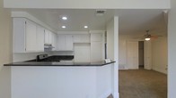 This single-story space showcases an open-concept layout that seamlessly connects the kitchen to the adjacent living area. The kitchen is outfitted with crisp white cabinetry that extends to the upper walls, offering generous storage. Dark granite countertops provide a striking contrast against the white surfaces, running along the perimeter and wrapping around a peninsula that serves as a natural divider between the kitchen and living space. A gas range and under-cabinet range hood are integrated into the design, and recessed lighting in the tray ceiling illuminates the kitchen efficiently. The peninsula's clean, curved edge adds a subtle architectural detail to the transition between spaces. The adjoining living area features neutral carpet flooring, providing a soft contrast to the hard surfaces in the kitchen. White walls throughout maintain a bright, cohesive aesthetic, while multiple ceiling vents suggest a well-distributed HVAC system. A door visible in the background hints at additional rooms or storage beyond the main living space. Overall, the layout is practical and functional, with a clean, understated aesthetic.