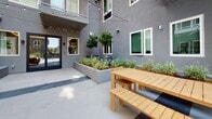 Located at 833 Fedora St in the heart of Los Angeles, this thoughtfully designed outdoor courtyard serves as a communal gathering space with a clean, contemporary aesthetic. Enclosed by smooth gray stucco walls and surrounded by the building's multi-story facade, the courtyard offers a sense of privacy while remaining open to the Southern California sky above.

The space is anchored by generously sized raised concrete planters filled with a curated mix of low shrubs, herbs, and neatly pruned trees, adding a vibrant touch of greenery that softens the modern architectural lines. String lights are strung overhead, creating an inviting ambiance suited for both daytime and evening use.

Two distinct seating areas define the layout. A lounge zone features solid teak-framed armchairs and a sectional with plush gray cushions and accent pillows, arranged around a low slatted coffee table — ideal for relaxed conversation. Adjacent to this, a dining area includes a teak picnic-style table with matching benches, offering a casual setting for outdoor meals or social gatherings.

The polished concrete flooring with subtle gray accent strips ties the space together cohesively. A set of dark-framed glass doors provides interior access, seamlessly connecting the outdoor courtyard to the building's interior. Overall, the courtyard exudes a refined yet welcoming character well-suited to urban community living.