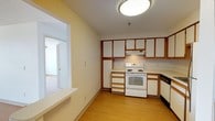 This single-floor residence offers two bedrooms and two bathrooms within a practical, well-organized layout. The kitchen serves as a central feature of the home, equipped with an electric range, dishwasher, and refrigerator. Cabinetry features white panels framed with warm wood trim, providing ample storage across both upper and lower cabinets. A built-in range hood sits above the stove, and light-toned countertops run along the perimeter, offering generous prep space. The kitchen floor is finished in light hardwood-style flooring, which continues into the adjacent living area, creating a cohesive flow throughout the main living spaces. A pass-through ledge connects the kitchen to the neighboring room, adding an open, communicative feel between spaces. The living area, visible through a wide doorway, is generously proportioned with neutral wall tones and carpeted flooring. The overall color palette is warm and understated, with cream-toned walls and warm wood accents throughout. Flush-mount ceiling lighting provides consistent illumination, and the layout, as seen in the floor plan, reflects an efficient and functional use of space suitable for everyday living.