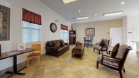 Located along Waterside Road in Prince George, Virginia, this single-story space is thoughtfully designed to serve as a functional community or professional gathering facility. The layout flows seamlessly across several distinct areas, each finished with consistent warm-toned tile flooring and crown molding throughout, contributing to a cohesive and polished aesthetic.

The main lounge area serves as the heart of the space, featuring multiple seating arrangements including a leather sofa, upholstered chairs, and a bistro-height table set. Natural light filters in through multiple windows dressed with striped valances, while recessed lighting and flush-mount fixtures ensure the space remains well-lit. French doors provide direct access to the exterior, enhancing the open, inviting atmosphere.

The conference room is equipped with a large oval wooden table surrounded by padded chairs, a wall-mounted flat-screen television, and recessed panel lighting overhead. Additional doors within the room lead to restroom facilities, making it well-suited for professional meetings or group gatherings.

The kitchen features warm oak cabinetry extending to the ceiling, a peninsula with a double sink and light countertops, and black appliances including a side-by-side refrigerator, range, and microwave. A large window above the peninsula floods the space with natural light, creating a bright and practical workspace ideal for event preparation or communal use.