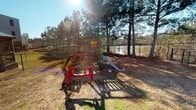 This outdoor recreational space offers a welcoming environment centered around a well-built wooden playground structure. The playset includes a raised platform, a slide, and red swing attachments, providing a variety of play options. The ground is covered with natural pine straw, giving the area a rustic, woodland character that blends seamlessly with the surrounding landscape.

The space is fully enclosed by a chain-link fence, ensuring a secure environment. Towering pine trees line the perimeter, offering partial shade and a natural canopy that filters sunlight throughout the day. A calm pond is visible just beyond the fencing, adding a peaceful, scenic backdrop to the play area.

The open layout of the yard allows for ample room to move freely around the playground structure. The combination of natural ground cover, mature trees, and the adjacent waterway creates a tranquil outdoor setting. Sunlight streams through the pine canopy, casting dappled shadows across the ground and enhancing the natural ambiance of the space. This outdoor area is well-suited for recreational use, offering both active play opportunities and a serene natural environment.