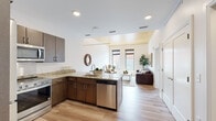 This contemporary single-floor residence showcases a thoughtfully designed open-concept layout that seamlessly connects the kitchen, dining, and living areas. The kitchen is fully equipped with dark espresso cabinetry, granite countertops, and a complete suite of stainless steel appliances, including a refrigerator, range, microwave, and dishwasher. Light hardwood flooring flows throughout, complementing the clean white walls and recessed lighting. A distinctive wood-paneled ceiling accent adds warmth and architectural interest to the main living area, which is bathed in natural light from large, floor-to-ceiling windows overlooking a lively urban streetscape. The dining area features a compact bistro-style table alongside bar seating at the kitchen peninsula, offering flexible options for casual meals. The living room is open and airy, with soaring wall height providing a generous sense of volume. The bedroom is a serene retreat, highlighted by a natural wood-slat ceiling, a tall vertical window with urban views, and light hardwood floors. The bathroom features large-format tile flooring, a glass-enclosed walk-in shower with white subway tile, and a double-sink vanity with dark cabinetry and white countertops. The overall ambiance is modern, clean, and urban.