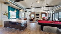 This dynamic single-floor space blends industrial-chic design with modern comfort, offering an engaging environment suited for socializing and community gathering. The open-concept layout is anchored by soaring ceilings with exposed ductwork, dark structural beams, and warm wood-look flooring that flows seamlessly throughout.

The welcoming reception area features large-format tile flooring, a sleek white front desk, exposed brick walls, and a cluster of pendant lights overhead. Staircase access and floor-to-ceiling windows flood the entry with natural light, framing views of the surrounding streetscape.

The expansive main lounge is a visual highlight, showcasing a full-size pool table with a bold red felt surface, a cozy seating area anchored by a rich leather sofa and vibrant multicolored area rug, and a fireplace set against a stone and brick surround with a wall-mounted TV above. Eclectic décor — including a starburst mirror, sculptural plants, and colorful accent chairs — adds personality and warmth.

A compact, well-appointed kitchenette features dark lower cabinetry, white countertops, open shelving, a beverage cooler, and a deep stainless steel sink, all bathed in natural light from a large picture window. Teal accent walls punctuate the space with bold color throughout, reinforcing its energetic, contemporary character.