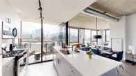 Located in Chicago's vibrant West Loop neighborhood, this contemporary unit at 160 N Elizabeth St showcases a striking industrial-modern aesthetic defined by exposed concrete ceilings, visible ductwork, and expansive floor-to-ceiling windows that flood every room with natural light. The open-concept living and kitchen area serves as the heart of the space, where a sleek white quartz island with bar seating anchors the kitchen, outfitted with stainless steel appliances, gray cabinetry, and track lighting. The living area flows seamlessly toward the floor-to-ceiling glazing, offering sweeping views of the Chicago skyline and access to an outdoor balcony. Each bedroom features large windows framing neighborhood rooftop views, exposed concrete ceilings, and hardwood-style flooring, creating a bright yet grounded atmosphere. The primary bedroom includes generous closet space and a refined, minimalist design. The bathroom is finished with large-format tile flooring, a clean floating vanity, and crisp white countertops. A hallway adorned with framed artwork connects the private and social areas of the unit, completing a cohesive, sophisticated living environment rooted in urban design.