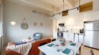This single-floor loft-style residence showcases an open-concept design that seamlessly integrates the living, dining, and kitchen areas into one cohesive space. Soaring ceilings with exposed structural beams and visible ductwork lend an industrial character, while light-toned hardwood flooring and a crisp white palette keep the atmosphere bright and contemporary. Track lighting runs overhead, adding both functional illumination and architectural interest.

The kitchen is outfitted with full stainless steel appliances — including a range, microwave, dishwasher, and refrigerator — complemented by white flat-front cabinetry, a subway tile backsplash, and clean countertops. It flows directly into the dining area, which features a marble-top table, and then into the living room, where large industrial-style windows frame urban cityscape views and fill the space with natural light.

The bedroom is generously proportioned with notably tall ceilings and elevated clerestory windows that allow additional natural light while maintaining privacy. Light hardwood flooring continues throughout. The bathroom features a frameless glass walk-in shower, tile flooring, a white vanity with stone-style countertop, and a round wall mirror. A hallway connects the private spaces, completing this well-designed, urban loft layout.