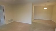 This single-floor space presents a straightforward and functional layout designed with simplicity and practicality in mind. The interior is finished in a neutral, warm beige color palette across the walls and ceiling, creating a calm and cohesive atmosphere throughout. Soft wall-to-wall carpeting in a complementary light tan tone covers the floors, adding warmth underfoot and contributing to the overall understated aesthetic.

The space features a partial wall partition that defines distinct zones within the open floor plan, allowing for a natural separation of areas while maintaining a sense of openness and connectivity between rooms. Clean white baseboards provide a crisp contrast to the neutral walls and carpeted floors. A floor-level ventilation register is visible, indicating a built-in climate control system integrated into the structure.

The layout flows seamlessly from one area to the next, with an open passage visible beyond the dividing wall, suggesting additional rooms or living areas. The overall ambiance is clean, minimal, and move-in ready, offering a blank canvas well-suited for a variety of interior design approaches. The space is practical and well-maintained, making it an accessible and versatile environment for everyday living.