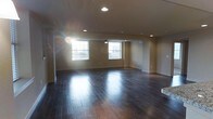 This single-floor space offers an open and airy layout designed for versatility and comfort. The most striking feature is the rich, dark hardwood flooring that stretches across the entire area, lending a warm and polished aesthetic that anchors the space. Recessed lighting is thoughtfully distributed across the ceiling, providing even, ambient illumination throughout. A ceiling fan is centrally positioned, adding both functionality and a subtle design element overhead. Large windows along one wall invite an abundance of natural light, creating a bright and welcoming atmosphere while offering views of the surrounding exterior. The neutral-toned walls complement the dark flooring, striking a classic contrast that keeps the space feeling balanced and clean. Smooth transitions between wall and floor are defined by crisp white baseboards, adding a refined finishing touch. The open floor plan provides generous room to move and allows for flexible arrangement, making the space well-suited for a variety of uses. The overall ambiance is one of understated elegance — clean, well-lit, and move-in ready with quality finishes throughout.