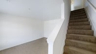 This two-story space presents a clean, move-in-ready environment defined by its neutral tones and straightforward layout. The main level features smooth tile flooring in a warm beige hue, complementing the crisp white walls and creating a bright, open atmosphere. The ceilings on this floor are smooth and well-proportioned, contributing to an airy feel throughout the space.

A staircase connects the two floors, finished with carpet in a muted taupe tone that adds a subtle warmth to the transition between levels. The staircase is framed by a white-painted banister and a classically styled newel post, lending a modest architectural detail to an otherwise minimalist interior. The stairwell benefits from natural light filtering in from above, suggesting a window on the upper level.

The overall design aesthetic is simple and understated, with a consistent white and neutral color scheme that provides a versatile backdrop for a range of interior styles. The space appears well-maintained and ready for occupancy, offering a blank canvas suitable for personalization.
