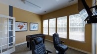This single-floor recreational space is designed with entertainment and leisure in mind, offering a welcoming environment for gaming and social activities. The room features warm golden-yellow walls that create an energetic yet inviting ambiance, complemented by carpeted flooring that adds a comfortable, casual feel underfoot.

Natural light floods the space through a row of large windows fitted with horizontal blinds, allowing for adjustable brightness throughout the day. The windows also provide pleasant views of the surrounding greenery outside.

The room is furnished with two ergonomic black gaming chairs, offering a dedicated space for video gaming or relaxed seating. A classic foosball table serves as a central recreational fixture, making the space ideal for group socializing and friendly competition. The walls are adorned with two framed graphic art pieces that add a modern, playful aesthetic to the room.

Overhead fluorescent lighting ensures the space remains well-lit even when natural light is limited. The overall layout is open and functional, with enough room to move comfortably between the various gaming and leisure stations. This thoughtfully arranged game room provides a fun and dynamic environment suited for recreation and community engagement.
