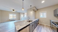 Located at 2067 11th Ave E in Menomonie, Wisconsin, this thoughtfully designed one-bedroom, one-bathroom unit offers a clean, modern aesthetic throughout. The open-concept kitchen and living area serves as the heart of the space, anchored by a large granite-topped island with an undermount sink and pendant lighting overhead. Gray shaker-style cabinetry lines the kitchen walls, complemented by a full suite of stainless steel appliances including a refrigerator, range, microwave, and dishwasher. Light-toned luxury vinyl plank flooring flows throughout the main living areas, enhancing the bright, airy feel created by multiple windows that invite natural light. The living room features a ceiling fan and an exterior view of the surrounding landscape. Carpeted stairs lead to additional areas of the unit. The bedroom is a comfortable retreat with plush carpet, a ceiling fan, and a large window. It connects to a generously sized walk-in closet outfitted with wire shelving on multiple walls, offering ample storage. The full bathroom includes a tub-shower combination, a gray vanity with light countertop, and matching hardwood-style flooring. Neutral wall tones and consistent modern finishes tie the entire space together cohesively.