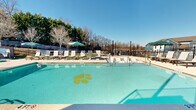 Located at 111 Cochran Rd in Clemson, SC, this outdoor amenity complex delivers a well-appointed recreational experience designed with community living in mind. At its centerpiece is a generously sized swimming pool featuring a distinctive Clemson tiger paw print on the pool floor — a nod to the local university spirit. The pool is surrounded by an expansive sun deck lined with white lounge chairs and shade umbrellas, providing ample space for relaxation under the open South Carolina sky. Depth markers and safety signage are clearly posted throughout the pool area, and a dedicated restroom facility is conveniently accessible poolside. Adjacent to the pool, a covered outdoor recreation area features corrugated metal roofing, warm string lighting, and exposed brick masonry walls, creating a casual yet inviting atmosphere. This space is equipped with a ping pong table and comfortable lounge seating, making it ideal for social gatherings. A separate lawn area showcases lush artificial turf, multiple picnic tables, and a built-in outdoor grilling station with dual grills — perfect for cookouts and outdoor dining. The entire complex is enclosed by a black iron fence, offering both security and an open, airy feel. Mature trees and greenery along the perimeter provide natural framing and shade throughout the grounds.