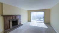 This single-floor space offers a welcoming and functional layout centered around a comfortable living area. The room is finished with soft wall-to-wall carpeting that adds warmth underfoot, while the light yellow walls lend a bright and airy atmosphere throughout.

A standout feature of the living area is the classic brick fireplace, complete with a dark wood mantel that serves as a natural focal point. Its traditional craftsmanship adds character and a sense of coziness to the space.

Natural light floods the interior through large sliding glass doors dressed with vertical blinds, which open directly onto a balcony. The outdoor access provides scenic views of the surrounding landscape, including hills and mature trees, creating a seamless connection between the interior and the outdoors.

The ceiling features a textured finish, adding subtle visual interest and depth to the room. Neutral tones throughout the space create a versatile backdrop well-suited to a variety of interior styles. Overall, the space balances comfort and functionality, with its fireplace, abundant natural light, and outdoor views contributing to a relaxed and inviting ambiance.