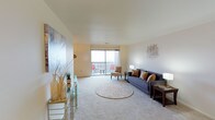 This single-floor residence centers around a spacious living area designed to maximize comfort and natural light. The room features wall-to-wall carpeting in a soft neutral tone, providing a warm and inviting foundation throughout the space. Large sliding glass doors dominate one wall, offering direct access to an outdoor balcony and flooding the interior with abundant natural light while framing open exterior views. The living area is generously proportioned, comfortably accommodating a full seating arrangement that includes a tufted sofa with accent pillows and a coordinating armchair. A round area rug anchors the central seating zone, adding texture and definition to the open layout. The walls are finished in a clean, light neutral tone, enhanced by decorative wall art that adds visual interest without overwhelming the space. A gold-toned accent console along one wall provides both style and functional display space. Vertical window treatments allow for flexible light control. The overall ambiance is bright, airy, and well-balanced, making the living area a comfortable and functional hub within the home.