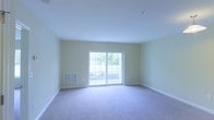 This single-floor space offers a clean and functional layout with a welcoming, light-filled atmosphere. The main living area is generously proportioned, featuring soft wall-to-wall carpeting in a neutral tone that adds warmth underfoot. The walls are painted in a soft, pale yellow-green hue, contributing to an airy and calm ambiance throughout.

A standout feature of the living area is the large sliding glass door, which floods the room with abundant natural light and provides direct access to an outdoor balcony. The balcony, visible through the glass, is framed by a white railing and overlooks a lush, green outdoor setting, making it an ideal spot for relaxation or fresh air.

The space is equipped with practical built-in features, including a wall-mounted HVAC vent for climate control, as well as multiple electrical outlets thoughtfully placed along the baseboards. The white baseboards and trim provide a crisp contrast against the soft wall tones, adding a polished, finished look to the interior.

The smooth, white ceiling is clean and unadorned, enhancing the sense of openness and height in the room. Overall, this space combines understated design with functional details, making it a bright and versatile environment suitable for a variety of uses.