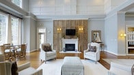 Located at 1600 Belle Point Dr in Mt. Pleasant, South Carolina, this single-story space offers a sophisticated and welcoming atmosphere inspired by coastal Lowcountry design. The main living area serves as the centerpiece of the space, anchored by a striking floor-to-ceiling wood-paneled accent wall that frames a classic white-mantled fireplace. Warm sconce lighting flanks a mounted television above the mantle, creating a balanced and functional focal point. The seating arrangement — composed of neutral-toned wingback chairs and a striped upholstered ottoman — is thoughtfully positioned to encourage conversation and relaxation. Underfoot, rich hardwood flooring extends throughout, complemented by a large area rug that defines the central seating zone. The elevated ceilings feature detailed coffered and paneled millwork finished in crisp white, lending a sense of grandeur and architectural refinement to the interior. Soft gray walls and abundant natural light from nearby windows contribute to an open, airy ambiance. The space transitions naturally toward an entry area with glass-paneled doors, reinforcing the connection to the outdoors. Overall, the design balances elegance with comfort, making it well-suited for communal use or upscale entertaining.