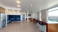 This single-floor space is designed as a contemporary communal lounge, combining multiple functional zones within one open, airy layout. The interior is finished with warm wood-look flooring and crisp white walls throughout, creating a clean, modern aesthetic complemented by recessed and flush-mount ceiling lighting.

The lounge area features a tufted cognac leather sofa paired with a sculptural wood side table, positioned on a textured area rug. Opposite, a wall-mounted flat-screen TV sits above a low-profile wood credenza with drawer storage. Two tall windows flood this zone with natural light and offer views of the surrounding cityscape.

A distinct seating nook is defined by a bold navy blue upholstered banquette with small round pedestal tables, ideal for intimate gatherings. A long wood conference-style dining table with modern black-and-gold chairs anchors the workspace or dining area nearby.

The kitchen includes light wood cabinetry, a marble-look countertop, glossy subway tile backsplash, and stainless steel appliances including a dishwasher, refrigerator, and microwave.

A standout feature is the expansive folding glass wall that opens the dining area directly onto a spacious outdoor terrace, offering sweeping open views and seamless indoor-outdoor flow.