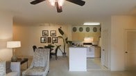 This single-floor residence offers an open-concept layout that effortlessly integrates the living, dining, and kitchen areas into one cohesive space. The living area is finished with soft carpeting underfoot and is illuminated by a ceiling fan with integrated lighting, lending both comfort and practicality. A set of framed wall art and a lush tropical houseplant add character and warmth to the space, while a dining table with chairs anchors the transition between the lounge and kitchen zones. Track lighting along the wall provides additional illumination flexibility throughout the shared area. The kitchen is defined by a peninsula-style breakfast bar that serves as a natural divider between the cooking and living spaces. Light-toned cabinetry lines the upper walls, complemented by under-cabinet lighting, and the kitchen is equipped with a white range and oven. Decorative plate accents are displayed above the upper cabinets, adding a homey touch. A door visible near the kitchen hints at additional utility or storage access. Overall, the space carries a cozy, welcoming ambiance with a practical, well-organized layout suited for everyday living.