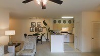 This single-floor residence offers an open-concept layout that effortlessly integrates the living, dining, and kitchen areas into one cohesive space. The living area is finished with soft carpeting underfoot and is illuminated by a ceiling fan with integrated lighting, lending both comfort and practicality. A set of framed wall art and a lush tropical houseplant add character and warmth to the space, while a dining table with chairs anchors the transition between the lounge and kitchen zones. Track lighting along the wall provides additional illumination flexibility throughout the shared area. The kitchen is defined by a peninsula-style breakfast bar that serves as a natural divider between the cooking and living spaces. Light-toned cabinetry lines the upper walls, complemented by under-cabinet lighting, and the kitchen is equipped with a white range and oven. Decorative plate accents are displayed above the upper cabinets, adding a homey touch. A door visible near the kitchen hints at additional utility or storage access. Overall, the space carries a cozy, welcoming ambiance with a practical, well-organized layout suited for everyday living.