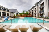 Located at 16200 Croft Drive in Charlotte, NC, this outdoor amenity space is designed to deliver a resort-style experience within a modern residential community. At the heart of the space is a generously sized swimming pool with crystal-clear turquoise water, framed by a wide pool deck furnished with rows of lounge chairs, perfect for relaxing and soaking up the Carolina sun. A shallow wading ledge with molded lounge seats provides an inviting entry point into the pool, adding a distinctive leisure feature to the area.

Surrounding the pool, the deck transitions into well-appointed seating zones, including shaded areas outfitted with patio umbrellas and additional chaise lounges. A pergola-style structure at one end of the pool area offers a covered gathering spot, complemented by lush greenery and landscaped planting beds that soften the hardscape throughout.

A separate courtyard area adjacent to the main pool features bistro-style table and chair sets amid young trees and manicured landscaping, providing a quieter outdoor retreat. The multi-story buildings enclosing the space feature a contemporary design with brick and board-and-batten exterior finishes, private balconies, and warm wood-accented soffits, all contributing to a cohesive and attractive outdoor environment ideal for community living.