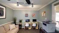 This single-floor space functions as a well-appointed business center and lounge, thoughtfully designed to support both productivity and comfort. The walls throughout are finished in a calming sage green, accented by white chair rail molding and crown molding that lend a polished, cohesive aesthetic.

The primary workstation area features two individual computer desks, each equipped with a monitor and a modern white chair, making it an ideal spot for focused work. Track lighting overhead, complemented by a ceiling fan, ensures the space is well-lit and comfortable. A printer and a filing station are also conveniently accessible. Decorative wall art with digital and typography themes adds a creative touch to the functional setup.

A cozy lounge area adjacent to the workstations includes a cushioned sofa with decorative pillows and a framed black-and-white photograph mounted above, creating a relaxed atmosphere for breaks or informal meetings. Built-in white shelving units provide practical storage alongside a secondary door for easy access.

A pass-through window connects to an adjoining room, which appears to serve as a refreshment and meeting area, featuring a coffee station, a dining or conference table with chairs, and an abundance of natural light from large windows. Patterned carpeting runs throughout, tying the spaces together with a warm, welcoming finish.