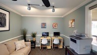 This single-floor space functions as a well-appointed business center and lounge, thoughtfully designed to support both productivity and comfort. The walls throughout are finished in a calming sage green, accented by white chair rail molding and crown molding that lend a polished, cohesive aesthetic.

The primary workstation area features two individual computer desks, each equipped with a monitor and a modern white chair, making it an ideal spot for focused work. Track lighting overhead, complemented by a ceiling fan, ensures the space is well-lit and comfortable. A printer and a filing station are also conveniently accessible. Decorative wall art with digital and typography themes adds a creative touch to the functional setup.

A cozy lounge area adjacent to the workstations includes a cushioned sofa with decorative pillows and a framed black-and-white photograph mounted above, creating a relaxed atmosphere for breaks or informal meetings. Built-in white shelving units provide practical storage alongside a secondary door for easy access.

A pass-through window connects to an adjoining room, which appears to serve as a refreshment and meeting area, featuring a coffee station, a dining or conference table with chairs, and an abundance of natural light from large windows. Patterned carpeting runs throughout, tying the spaces together with a warm, welcoming finish.