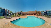 This single-story clubhouse serves as the social and administrative hub of a residential community, offering a well-appointed interior complemented by impressive outdoor amenities. Outside, a generously sized freeform swimming pool takes center stage, surrounded by a stone-tile sundeck lined with lounge chairs. A covered outdoor bar area and lush green lawn sit adjacent to the pool, all enclosed within a wrought iron fence for privacy and security.

Inside, the open-plan interior is bright and welcoming, featuring light wood-look flooring, vaulted ceilings with ceiling fans, and an abundance of natural light from large windows overlooking the pool area. The lounge area anchors the space with a stone fireplace, wall-mounted television, and a seating arrangement of plush navy blue accent chairs accented with yellow throw pillows. A leasing or business center desk area with computer workstations is seamlessly integrated into the open layout.

Toward the back, a casual dining area offers round white tables paired with vibrant yellow chairs, creating a cheerful, modern aesthetic. A kitchenette with white cabinetry, a decorative mosaic tile backsplash, open shelving, and a coffee station provides refreshment convenience. The cohesive blue and yellow color palette, combined with contemporary artwork and clean white walls, gives the entire space a fresh, energetic, and community-focused atmosphere.