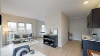 This single-floor studio is thoughtfully designed with a contemporary open-concept layout that maximizes functionality and flow. Light hardwood flooring runs throughout the entire unit, lending a clean, cohesive aesthetic to every area.

The main living space is bright and airy, benefiting from an abundance of natural light delivered through large windows and sliding glass doors that open directly onto a private balcony. The balcony offers outdoor views of the surrounding neighborhood and greenery. A half-wall partition subtly divides the living and sleeping areas while maintaining an open, connected feel.

The kitchenette is compact yet well-appointed, featuring dark cabinetry with upper and lower storage, light countertops, a single-basin sink, a built-in microwave, and additional appliances. It connects seamlessly to the dining area, which is positioned to take full advantage of the large windows and natural light.

The sleeping area is neatly tucked behind the partition, providing a sense of privacy within the open floor plan. A dedicated bathroom includes a wall-mounted sink, toilet, and a mirrored medicine cabinet, finished in neutral tones for a clean, minimal look.

Overall, the unit presents a modern, minimalist design characterized by neutral warm tones, efficient use of space, and a strong connection to natural light and outdoor views.