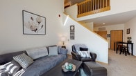 Located at 2707 Park Place Ln in Janesville, Wisconsin, this two-story space showcases a thoughtfully designed layout with standout architectural details across both levels. The main living area commands attention with soaring double-height ceilings, creating an open and airy atmosphere. A wood-railed staircase leads to the upper level, adding warmth and visual interest to the space. The living room is generously proportioned, offering ample room for comfortable seating arrangements, and benefits from natural light that enhances the bright, neutral color palette used throughout. A secondary sitting or flex space on the main level features a large window that invites additional natural light and provides versatile functionality. The upper level opens to a spacious loft-style bedroom with plush carpeting, providing a serene and private retreat. A corner entertainment area with built-in shelving adds practical character to the sleeping space. The bathrooms are well-appointed, with one featuring warm wood cabinetry, tile flooring, a large vanity mirror with strip lighting, and clean, classic finishes. A second bathroom includes a tiled tub and shower combination. Throughout the space, neutral walls, carpeted floors, and warm wood trim create a cohesive and welcoming ambiance.