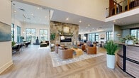 Located at 83 Terrace Greene Dr in Barboursville, VA, this generously sized single-floor community space is a masterclass in modern transitional design. The layout flows seamlessly across multiple distinct lounge and gathering areas, all unified by warm-toned hardwood flooring — including a striking herringbone pattern near the entry — and crisp white walls that amplify the sense of openness.

At the heart of the space stands a towering natural stone fireplace, serving as a dramatic focal point around which leather and upholstered seating arrangements invite relaxed conversation. Soaring double-height ceilings in key areas are complemented by expansive black-framed windows that flood the interior with natural light and frame outdoor views of the surrounding community.

The reception area features a curved wood desk beneath a double-ring chandelier, with a grand staircase and floor-to-ceiling glazing creating an impressive, light-filled entrance. Throughout the space, carefully curated seating vignettes — from plush sectionals and swivel chairs to banquette-style bench seating with bistro tables — offer varied settings for lounging, dining, or working. A cozy vestibule with sage-green shiplap walls and terracotta velvet chairs provides a welcoming transitional moment. Lush indoor plants and layered area rugs add warmth and texture throughout.
