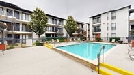 Located at 15678 Knoll Trail Dr in Dallas, TX, this outdoor amenity area is the central gathering space of a multi-story residential apartment community. The focal point of the space is a large, rectangular swimming pool with clear turquoise water, framed by a generous concrete sundeck. The deck is outfitted with multiple lounge chairs and colorful patio umbrellas, providing ample space for sunbathing and relaxation. The pool area is enclosed by a wrought iron fence, ensuring both safety and a defined boundary for the amenity space. Surrounding the pool on multiple sides are the community's three-story residential buildings, finished in a neutral stucco exterior with dark-trimmed balconies and exterior stairwells, accented by mature magnolia and shade trees that add a natural, welcoming element to the courtyard setting. Adjacent to the pool deck is a covered outdoor pavilion supported by bold red columns and a dark wood pergola-style roof. This entertaining area includes built-in grilling stations constructed with stone detailing, as well as bench and lounge seating, making it an ideal space for social gatherings. The overall ambiance is that of a well-appointed residential community amenity, blending functional outdoor living with comfortable communal spaces in the heart of Dallas.