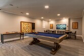 This single-floor amenity space is thoughtfully designed to provide residents with a variety of social and recreational options. At its heart is a dedicated game room, clearly marked with signage, that houses a full-size pool table with a rich wood frame and blue felt surface, complemented by a wall-mounted Scrabble board. Adjacent to the billiards area, two round card tables are surrounded by plaid upholstered armchairs, offering comfortable seating for card games or casual gatherings. A flat-screen television is mounted on the wall, and decorative artwork — including backgammon boards and framed pieces — adds character to the room. The light wood-tone flooring and recessed lighting throughout give the space a clean, modern aesthetic with warm crown molding details. A welcoming foyer area with lounge chairs and a marble-top side table serves as a transition point into the game room corridor. The broader facility opens into a dramatic multi-level common area featuring soaring ceilings, an elegant geometric chandelier, a patterned area rug with circular seating arrangements, a built-in bookcase, and an abundance of natural light from large windows — creating an inviting and vibrant communal hub.