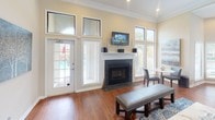 This well-appointed single-floor space exudes a warm, inviting ambiance anchored by rich hardwood flooring that flows seamlessly throughout. The main living area is generously proportioned, featuring a classic white-mantled fireplace with dark slate tile surround, above which a wall-mounted flat-screen television is positioned for comfortable viewing. Crown molding details add a refined architectural touch to the warm beige walls.

A striking bay window area with an arched transom creates a bright, sunlit nook perfect for seating or a workspace, fitted with a ceiling fan and recessed lighting. Tall windows with white plantation blinds line the perimeter, flooding the space with natural light and offering views of an outdoor area, including a glimpse of a pool beyond the French door.

The open layout connects the main seating area to a hallway corridor, framed by a wide archway and accented with gallery-style framed artwork. A dark wood credenza along one wall provides both storage and display space, complemented by a decorative round mirror. The seating arrangement — featuring neutral upholstered chairs and a patterned area rug in blue tones — contributes to the space's polished yet comfortable character. High transoms above the windows and doors further enhance the sense of openness and volume throughout.