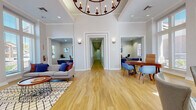 This single-floor community or leasing office is thoughtfully designed to be both welcoming and functional. The main lobby serves as the heart of the space, featuring soaring ceilings, crown molding, and wide-plank light wood flooring that runs throughout. A striking circular chandelier with candle-style lighting anchors the central entry area, adding a touch of elegance overhead.

Large floor-to-ceiling windows along multiple walls flood the interior with abundant natural light, offering views of the surrounding outdoor landscape. Multiple distinct seating areas are arranged throughout the open layout — including a casual lounge with a modern gray sofa, patterned area rug, and colorful accent pillows, as well as groupings of leather and upholstered chairs for informal conversation.

A dedicated private office area features a rich wood desk paired with blue upholstered guest chairs, ideal for one-on-one consultations. White cabinetry with light countertops provides additional workspace and storage. A long hallway extends from the main lobby, connecting to further rooms and amenities within the facility.

Double glass entry doors with transom windows create a bright, open threshold to the outdoors. The overall ambiance is polished yet approachable, combining contemporary furnishings with classic architectural details for a professional and inviting environment.