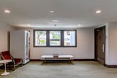 Located at 222 Columbia St in Santa Cruz, California, this single-floor common area serves as a welcoming communal hub. The space is composed of a central living/lounge area and two connecting hallways, all finished with patterned wall-to-wall carpeting and clean white walls accented by dark wood baseboards.

The lounge area is thoughtfully furnished with mid-century-inspired leather lounge chairs in warm rust tones, paired with a round pedestal side table — ideal for casual conversation or relaxation. A tall potted monstera plant adds a lush, natural element, while a black-and-white photographic print of a surfer on the beach lends a distinctly Santa Cruz character to the space. A vending machine provides added convenience for occupants.

A built-in wall-mounted desk with a cushioned rolling chair and a vine-draped mirror creates a functional work nook at the hallway entrance. Recessed ceiling lighting throughout ensures a bright, evenly lit atmosphere, complemented by natural light streaming through dark-framed windows that offer views of surrounding greenery.

The hallways are wide and well-lit, lined with doors and framed artwork, extending the clean and organized aesthetic of the lounge. Overall, the space strikes a balance between practical functionality and a relaxed, coastal-inspired ambiance reflective of its Santa Cruz setting.