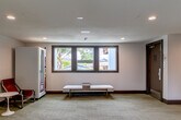 Located at 222 Columbia St in Santa Cruz, California, this single-floor common area serves as a welcoming communal hub. The space is composed of a central living/lounge area and two connecting hallways, all finished with patterned wall-to-wall carpeting and clean white walls accented by dark wood baseboards.

The lounge area is thoughtfully furnished with mid-century-inspired leather lounge chairs in warm rust tones, paired with a round pedestal side table — ideal for casual conversation or relaxation. A tall potted monstera plant adds a lush, natural element, while a black-and-white photographic print of a surfer on the beach lends a distinctly Santa Cruz character to the space. A vending machine provides added convenience for occupants.

A built-in wall-mounted desk with a cushioned rolling chair and a vine-draped mirror creates a functional work nook at the hallway entrance. Recessed ceiling lighting throughout ensures a bright, evenly lit atmosphere, complemented by natural light streaming through dark-framed windows that offer views of surrounding greenery.

The hallways are wide and well-lit, lined with doors and framed artwork, extending the clean and organized aesthetic of the lounge. Overall, the space strikes a balance between practical functionality and a relaxed, coastal-inspired ambiance reflective of its Santa Cruz setting.