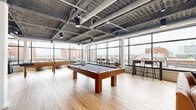 Located at 628 Ellicott Street in Buffalo, NY, this generously proportioned amenity lounge offers a dynamic blend of industrial design and modern comfort. The open-plan layout spans a single floor, providing an expansive, flexible space suited for both social gatherings and relaxed recreation. Exposed concrete ceilings with visible ductwork and pendant lighting give the space a distinctly urban, loft-inspired aesthetic, while light wood-plank flooring adds warmth throughout.

Floor-to-ceiling windows wrap along the perimeter, flooding the interior with abundant natural light and framing sweeping views of the Buffalo skyline and surrounding brick architecture. A central billiards table anchors the recreation area, complemented by a shuffleboard table and a high-top bar seating zone. The lounge area features a modern gas fireplace set against a boldly patterned accent wall, surrounded by plush sofas, accent chairs, and vibrant area rugs that add a pop of color and personality.

A sleek kitchenette with a stainless steel counter, mosaic tile backsplash, open shelving, and bar-height seating provides a functional social hub. Exposed brick walls weave throughout the space, nodding to the building's historic character. Direct access to an outdoor terrace with city views further enhances the appeal of this well-appointed communal space.