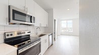 Located on E 23rd Ave in Aurora, Colorado, this single-floor unit showcases a clean, modern design throughout. The open-concept layout connects the kitchen and living area seamlessly, creating a bright and functional living environment. Light hardwood-style flooring runs continuously throughout the space, contributing to the unit's cohesive, contemporary aesthetic.

The kitchen is well-appointed with white flat-front cabinetry, stainless steel appliances including a refrigerator, electric range, built-in microwave, and dishwasher. A white subway tile backsplash adds a classic touch against the crisp white surfaces, while recessed lighting ensures the area remains well-lit.

The living area benefits from a large picture window that invites generous natural light and offers expansive views of the surrounding Aurora cityscape. The bedroom continues the bright, minimal theme with white walls, light flooring, a sizable closet, and its own large window framing additional outdoor views.

The bathroom features dark vertically-stacked tile in the shower enclosure, contrasting cleanly with the white walls and light flooring. Overall, the space presents a fresh, move-in-ready environment with a consistent modern design language throughout.