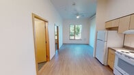 Located at 9715 NE Glisan St in Portland, Oregon, this single-floor unit offers a clean, modern design with a straightforward, functional layout. The main living area is notably open and airy, benefiting from tall ceilings that amplify the sense of space. Light-toned hardwood-style flooring runs throughout, contributing to a bright, cohesive aesthetic. A ceiling fan is centrally positioned overhead, providing both airflow and ambient lighting. A window overlooking a railed outdoor area frames views of surrounding greenery, allowing natural light to fill the room. A sliding door panel provides a flexible room division option within the living space.

The kitchen is efficiently arranged along one wall and features light wood cabinetry — both upper and lower — with neutral-toned countertops, a stainless steel sink, a white electric range, and a full-size refrigerator. Under-cabinet lighting adds practicality to the workspace. Ample storage is provided through the generous cabinet configuration.

The bathroom maintains the same clean, contemporary style, with light wood vanity cabinetry, a gray countertop, a single sink, and a full shower-tub combination. White walls and simple chrome fixtures complete the space's understated design.