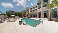 Located at 211 River Landing Dr in Charleston, SC, this amenity space combines classic Southern architecture with resort-style outdoor living. The building's distinctive red brick exterior, accented by teal-framed windows and arched entryways, sets an elegant tone throughout. Towering brick columns and high ceilings define the covered outdoor pavilion, which features lush vertical green walls that bring a vibrant, garden-like atmosphere indoors. The pavilion is furnished with wicker seating dressed in navy blue cushions, complemented by woven area rugs and glass-topped side tables, creating a comfortable and stylish lounge environment. Dramatic white drape curtains frame open views directly out to the pool area, allowing natural light to flood the space. A large flat-screen television is mounted on a greenery-covered wall, making the pavilion ideal for social gatherings. The adjoining outdoor pool area is expansive, featuring crystal-clear water, a wading shelf, and a generous sun deck lined with lounge chairs and shade umbrellas. Mature palm trees and surrounding landscaping enhance the resort ambiance. Brick-paved walkways connect the interior lounge to the pool deck, and a dining nook with bistro-style seating is visible through large glass doors, completing this well-appointed amenity offering.