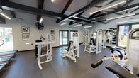 Located at 9961 Lurline Ave in the Chatsworth neighborhood of Los Angeles, this single-floor fitness center is thoughtfully designed to support a comprehensive workout experience. The space is divided into distinct training zones, including a strength equipment area and a dedicated cardio section, each outfitted with a broad selection of modern machines.

The strength zone features multiple white cable and weight machines, benches, and a leg roller, while the cardio area is equipped with elliptical trainers, stationary bikes, and stability balls for functional training. Full-length mirrors line the walls throughout, enhancing the sense of openness and allowing users to monitor their form.

Architecturally, the space is distinguished by its dramatic vaulted ceiling with exposed dark wooden beams set against white-paneled surfaces, lending an industrial-meets-contemporary aesthetic. Overhead linear lighting provides bright, even illumination throughout. The durable, dark rubber flooring is well-suited to heavy equipment and high-traffic use.

Large glass double doors on multiple sides of the facility invite natural light and offer views of the surrounding landscaped grounds and residential buildings. Wall-mounted fans and mounted flat-screen televisions add to user comfort. Framed artwork accents the walls, contributing a polished, welcoming atmosphere to this well-appointed fitness facility.