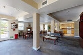 Located at 399 East Ct in San Jose, CA, this single-floor community space offers a generous, open layout designed to accommodate a variety of group activities and social gatherings. The main area features a expansive dining room furnished with multiple long wooden tables and chairs, providing ample seating for large groups. Structural columns define zones within the open floor plan while maintaining a sense of connectivity throughout the space. A decorative tiled fireplace serves as a focal point in the dining area, flanked by windows that allow natural light to brighten the room. The warm beige walls and plush carpeted flooring contribute to a welcoming, comfortable atmosphere. Recessed ceiling lighting ensures the space is well-lit throughout. A separate lounge area with a glass-top coffee table and bench seating offers a more relaxed setting. The fully equipped kitchen features warm wood cabinetry, tile countertops, a full-size refrigerator, stove, microwave, and dishwasher, all set on easy-to-clean tile flooring. A pass-through counter connects the kitchen to the main gathering area, making it convenient for serving and socializing. Overall, the space is thoughtfully arranged to support both casual interaction and organized community events.