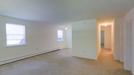 This single-floor space presents a clean, straightforward layout centered around a generously sized open living area. The room is finished with neutral white walls and wall-to-wall carpet in a warm beige tone, creating a blank canvas well-suited for a variety of furnishing arrangements. A baseboard heater runs along the exterior wall beneath a standard window, which allows natural light to filter into the space. The ceiling is smooth and white, fitted with a simple ceiling-mounted light fixture that provides general illumination to the main area.

A partial dividing wall separates the main living zone from an adjoining area, adding a degree of architectural definition without fully enclosing the space. This layout allows for a natural flow between areas while maintaining a sense of openness throughout. A hallway extends toward the rear of the unit, suggesting additional rooms beyond the main living space, with what appears to be a baseboard heater visible further down the corridor, indicating consistent heating throughout.

Overall, the space offers a practical, no-frills interior with a light, airy feel supported by its white walls, neutral flooring, and simple finishes.