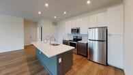 Located at 3900 W Broad St in Richmond, Virginia, this single-floor unit offers a well-designed layout with modern finishes throughout. Wood-style flooring flows consistently across the living areas and bedrooms, providing a warm, cohesive aesthetic.

The kitchen serves as a central highlight, featuring a large island with a white quartz countertop and a striking blue-gray base, stainless steel appliances including a range, microwave, and refrigerator, tall white upper cabinetry, dark lower cabinets, and a herringbone-patterned tile backsplash. Recessed lighting illuminates the space effectively.

The living and bedroom areas are generously sized, with multiple large windows dressed with horizontal blinds that invite ample natural light and offer views of the surrounding neighborhood. The layout, as seen in the 3D floor plan, demonstrates a spacious, open flow between the primary living areas.

One bedroom features a sliding barn-style door leading to a walk-in closet with built-in shelving. The bathrooms include clean, neutral tile flooring, white quartz vanity tops, dark wood cabinetry, large mirrors, and brushed nickel fixtures. One bathroom offers a full tub-shower combination, while another presents a vanity with a tall storage cabinet alongside.