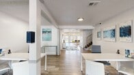 Located at 2700 Indian Creek Blvd in Oklahoma City, Oklahoma, this well-appointed single-floor space offers a generous and versatile layout designed for comfortable gathering and functionality. The interior is characterized by wood-look flooring that flows seamlessly throughout, bright white walls, and a clean, modern aesthetic accented with coastal-inspired artwork and decor.

The expansive living room is a standout feature, boasting a dramatic floor-to-ceiling white stone fireplace that serves as a striking focal point. Large multi-pane windows and glass French doors flood the room with natural light while offering views of the outdoor surroundings. A ceiling fan overhead enhances comfort in this open, airy space.

An open entry and corridor connect to a well-equipped kitchen featuring light blue cabinetry, ample counter space, a full-size refrigerator, dishwasher, microwave, and sink — ideal for hosting or everyday use. Adjacent to the kitchen is a bright dining nook with direct outdoor access through large windows and a glass door, providing a pleasant connection to the exterior grounds.

Additional open areas accommodate workstations and flexible-use seating, supported by abundant windows that maintain a light, welcoming atmosphere. A staircase with modern metal railings is also present within the space.