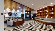 This thoughtfully designed amenity floor showcases a cohesive mid-century modern aesthetic throughout its various interconnected zones. The main corridor and central lounge immediately impress with a striking black-and-white geometric patterned floor, warm wood-paneled ceilings, and globe pendant lighting that set a sophisticated tone from the moment you enter.

A dramatic feature wall composed of diamond-shaped wood panels interspersed with mirrors and live moss panels serves as a focal point in one of the primary lounge areas, where teal velvet armchairs surround a marble coffee table atop a bold graphic rug. Built-in wood shelving units with curated décor accents appear throughout, adding warmth and visual interest.

The social kitchen and bar area features dark stone countertops, rich walnut cabinetry, white painted brick walls, open box shelving, and a large island with seating — ideal for casual gatherings. Adjacent is a cozy lounge nook with deep blue leather sectional seating, a patterned area rug, and a built-in book display along textured wall panels.

A dedicated game room offers a full-size pool table, intimate seating, and curated artwork. A bright café-style dining area with floor-to-ceiling windows floods the space with natural light and outdoor views, completing this well-appointed amenity level.