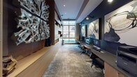 This contemporary co-working space is thoughtfully designed to support both focused work and creative inspiration. The elongated layout is anchored by a built-in workstation along one wall, equipped with multiple computer monitors and ergonomic seating, set against a rich, deep-teal accent wall adorned with large-scale abstract artwork. Directional wall-mounted lighting provides targeted illumination for each workstation.

Opposite the workstations, floor-to-ceiling built-in shelving in warm wood tones lines the wall, offering open display niches that house curated decorative objects and books, adding a sense of depth and sophistication. A striking sculptural wall piece serves as a dramatic focal point within the shelving system.

A large picture window at the far end of the room floods the space with natural light and offers views of the surrounding exterior, creating a bright visual terminus to the room. Adjacent to the window, a glass-enclosed private nook provides a quieter retreat furnished with a lounge chair — ideal for reading or informal meetings.

Textured, neutral-toned carpet spans the length of the room, softening the overall aesthetic and contributing to acoustic comfort. Recessed ceiling lighting complements the layered lighting scheme throughout. A built-in cabinetry unit along one end accommodates office equipment such as a printer, ensuring full functionality within the space.