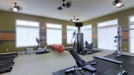 This fitness center offers a comprehensive workout environment designed to accommodate a wide range of exercise routines. The space is bathed in natural light, thanks to large windows with white blinds lining the walls, creating a bright and motivating atmosphere. The walls are painted in a warm olive green accented with horizontal orange and blue stripes, lending the space a dynamic and energetic character. Ceiling-mounted dual-fan fixtures provide ventilation and airflow throughout the room, contributing to comfort during workouts. The flooring transitions between protective rubber mats and a neutral carpet, helping to define distinct workout zones. Equipment includes a stationary exercise bike, a multi-station cable and weight machine, a leg press or extension machine, a weight bench, and several large stability balls, offering options for both cardio and strength training. A wall-mounted flat-screen television is positioned for easy viewing during exercise sessions. The overall layout is thoughtfully organized to maximize usable floor space while keeping equipment accessible and functional.