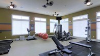 This fitness center offers a comprehensive workout environment designed to accommodate a wide range of exercise routines. The space is bathed in natural light, thanks to large windows with white blinds lining the walls, creating a bright and motivating atmosphere. The walls are painted in a warm olive green accented with horizontal orange and blue stripes, lending the space a dynamic and energetic character. Ceiling-mounted dual-fan fixtures provide ventilation and airflow throughout the room, contributing to comfort during workouts. The flooring transitions between protective rubber mats and a neutral carpet, helping to define distinct workout zones. Equipment includes a stationary exercise bike, a multi-station cable and weight machine, a leg press or extension machine, a weight bench, and several large stability balls, offering options for both cardio and strength training. A wall-mounted flat-screen television is positioned for easy viewing during exercise sessions. The overall layout is thoughtfully organized to maximize usable floor space while keeping equipment accessible and functional.