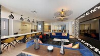 Located at 201 W Glenn Ave in Auburn, Alabama, this contemporary community lounge offers a thoughtfully designed, open single-floor layout that balances social gathering and relaxed lounging. Light-toned hardwood-style flooring runs throughout the space, complementing the crisp white and cool blue-gray walls and lending a bright, welcoming ambiance.

The living area serves as the centerpiece, anchored by a large flat-screen television mounted within a striking black-and-white geometric patterned accent wall. Bold, tufted navy blue sofas and accent chairs with orange and yellow pillows create vibrant, comfortable seating clusters, while patterned area rugs define distinct zones within the open floor plan. A ceiling-mounted projector adds an entertainment dimension to the space.

Distinctive black-and-white textured columns divide the lounge from a long communal bar-height counter lined with modern amber-hued chairs, ideal for individual work or group gatherings. Pendant lighting with sculptural, geometric fixtures adds dramatic visual interest overhead.

Toward the perimeter, a shuffleboard game table offers recreational appeal, and large windows and glass doors at one end of the room invite natural light and provide views of the surrounding area. Curated framed artwork adorns the walls, adding color and personality. The overall design is modern, energetic, and well-appointed for community living.